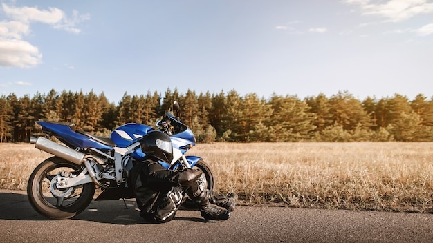 Biker in leather outfit and a black helmet sits on the asphalt road next to a sports motorcycle and looks into the distance