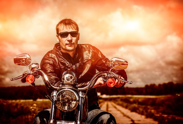 Foto uomo del motociclista che indossa una giacca di pelle e occhiali da sole seduto sulla sua moto guardando il tramonto.