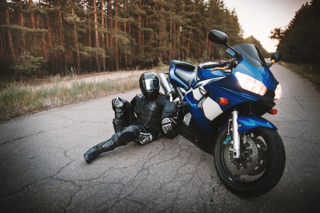 防護服と黒いヘルメットをかぶったバイカーは、道路上のバイクの隣に座っています。オートバイの近くで休んでいるモーターサイクリスト