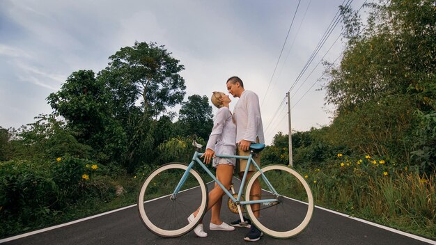 Biking road trip love couple on blue bike in white clothes on\
forest road just married woman and man kiss hugs stand on bicycle\
wedding honeymoon cycling cycle fix asia thailand ride tourism