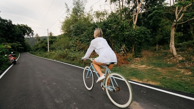 サイクリングロードトリップ。林道で白い服を着た青いバイクの女性。女の子は自転車に乗る。サイクリングサイクルの修正。アジアタイは観光に乗る。