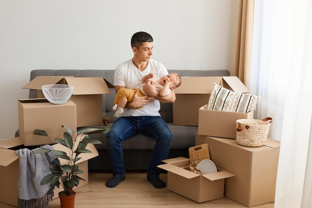 Binnen schot van aantrekkelijke brunette man met wit overhemd en spijkerbroek zittend op de bank met huilende peuter baby dochter slaperig baby kind moe tijdens verhuizing