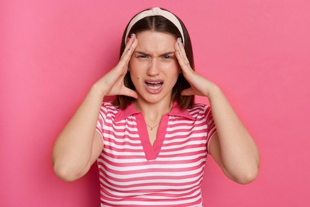 Foto binnen schot van geïrriteerd boos agressief jong meisje met haarband met gestreept casual t-shirt staande geïsoleerde roze achtergrond schreeuwend van boos met hoofdpijn