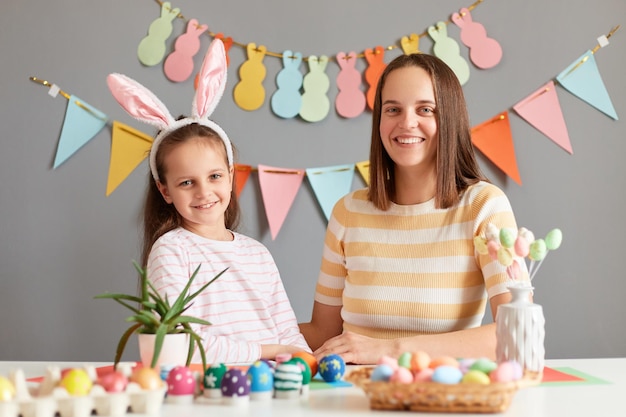 Binnen schot van glimlachende aantrekkelijke moeder en haar schattige dochter zittend aan tafel tussen gekleurde paaseieren gelukkige familie die zich voorbereidt op Pasen klein kind meisje met konijnenoren