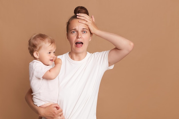 Binnen schot van jonge vermoeide geschokte moeder die een kind in haar armen houdt, een vrouw draagt een witte casual stijl Tshirt poseren met een baby geïsoleerd op bruine achtergrond schreeuwen houdt de hand op het hoofd