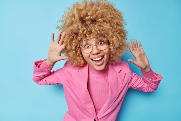 Binnen schot van positief goed geklede vrouw met krullend haar houdt handpalmen omhoog glimlacht breed heeft vrolijke stemming speelse uitdrukking draagt optische bril geïsoleerd over blauwe achtergrond kijk naar mij