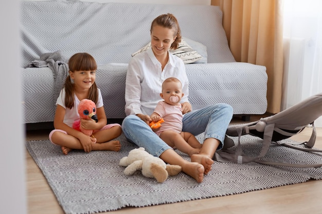 Binnen schot van schattige baby oudere zus en jonge moeder thuis zittend op de vloer vrouw met wit overhemd en spijkerbroek spelen met haar kinderen tijdens het weekend