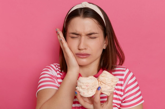 Binnen schot van trieste ongezonde vrouw met gestreept t-shirt poseren geïsoleerd over roze achtergrond voelt pijn in de tanden na het eten van zoete marshmallows hand op de wang houden