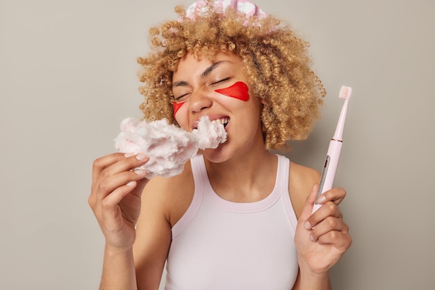 Binnen schot van vrouw met krullend haar bijt zoete suikerspin schadelijk voor tanden houdt elektrische borstel past rode schoonheidspleisters draagt casual t-shirt geïsoleerd over grijze achtergrond ongezond voedsel