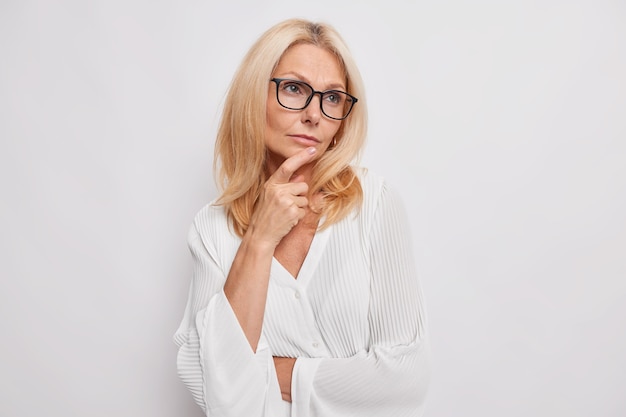 Binnenopname van doordachte lichtharige vrouw van middelbare leeftijd herinnert zich iets uit het leven kijkt weg houdt hand op kin bedenkt plan draagt bril voor zichtcorrectie stijlvolle zijden witte blouse