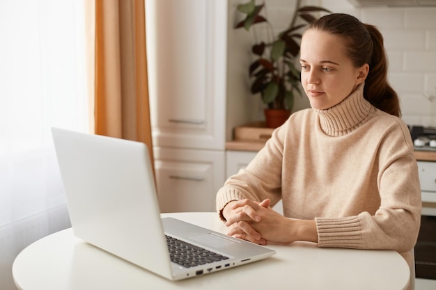 Binnenopname van een aantrekkelijke rustige jonge vrouw met een beige trui die in de keuken zit met een notebook aan het werk met een online college en kijkt naar de laptop met een serieuze uitdrukking