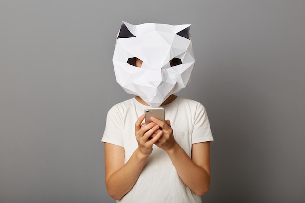 Binnenopname van een anonieme vrouw met een kattenpapieren masker en een wit T-shirt poseren geïsoleerd over een grijze achtergrond met behulp van een mobiele telefoon die berichten in sociale netwerken beantwoordt