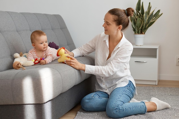 Binnenopname van een blanke vrouw met een wit overhemd en een spijkerbroek die op de vloer zit en tijd doorbrengt met de moeder van een dochtertje met een knotkapsel dat speelgoed aan het kind laat zien