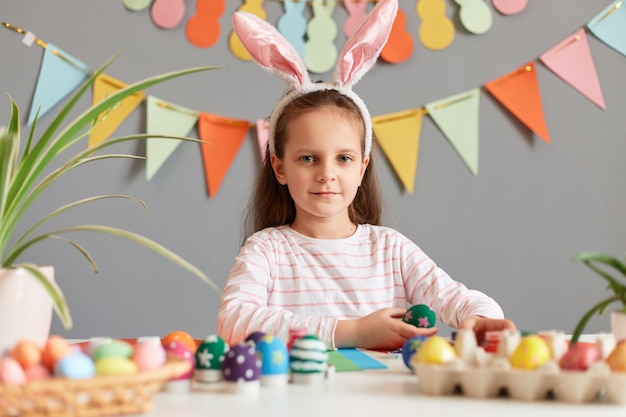 Binnenopname van een bruinharig meisje gekleed in konijnenoren die aan tafel zitten en naar de camera kijken die zich voorbereidt op Pasen zelf eieren schilderen tegen een versierde grijze muur
