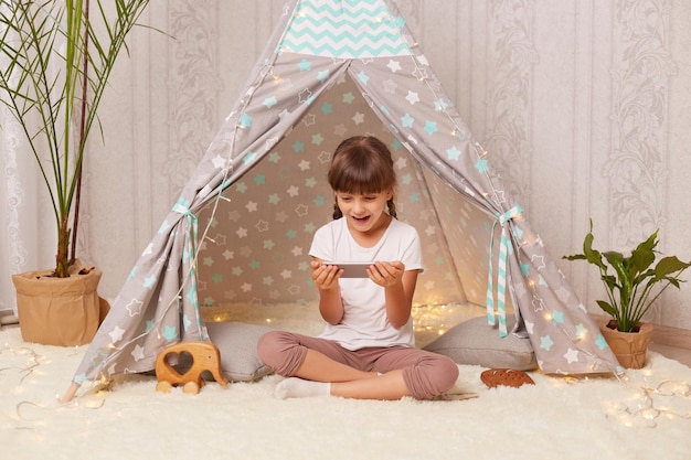 Binnenopname van een geschokt meisje met staartjes met een wit t-shirt in een wigwam thuis en met behulp van een telefoon kijkend naar het display met open mond ziet iets verbazingwekkends op internet