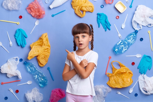 Binnenopname van een geschokt, verdrietig meisje met een casual T-shirt kijkt droevig naar de camera die alle plastic afval laat zien dat bezorgd is over een ernstig milieuprobleem dat zich voordoet tegen een blauwe achtergrond