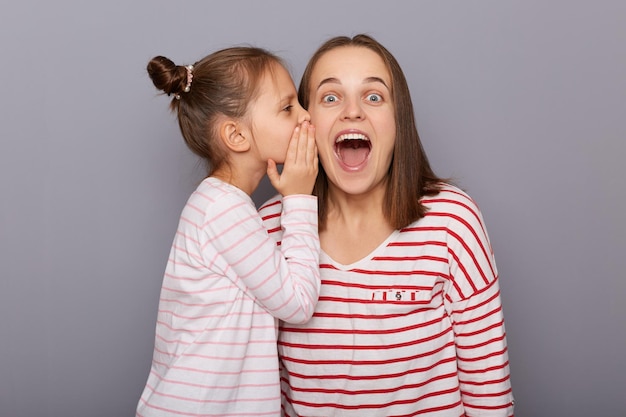 Binnenopname van een geschokte vrouw die met open mond naar de camera kijkt klein meisje met haarbroodjes die geheim zeggen tegen mama hoor familie dragen casual kleding staande geïsoleerd over grijze achtergrond