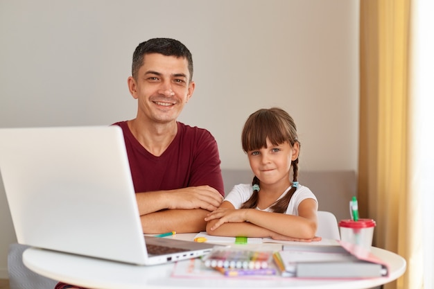 Binnenopname van een glimlachende knappe man die aan tafel zit met zijn dochtertje voor een laptop, kijkend naar de camera met een vrolijke uitdrukking, afstandelijk onderwijs.