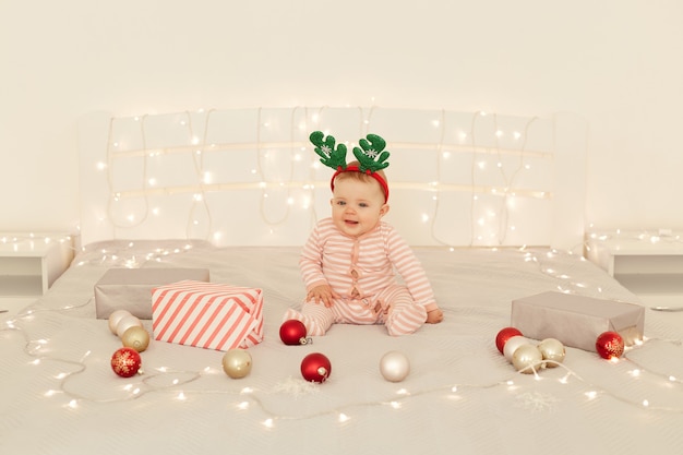 Binnenopname van een klein charmant meisje met een sleeper en hertenhoorns carnaval hoepel zittend op bed in een lichte kamer omringd met kerstversiering.