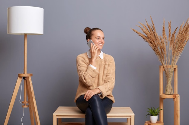 Binnenopname van een mooie blanke vrouw met een knotkapsel in beige zittend op een stoel en pratend op een smartphone terwijl ze opzij kijkt met positieve emoties die haar vriend belt
