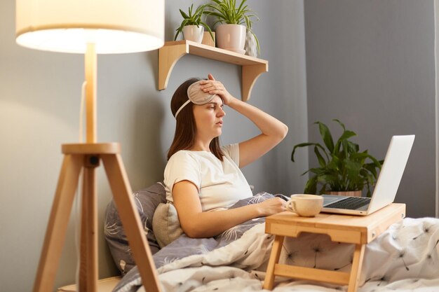 Binnenopname van een ongezonde zieke vrouw met een slaapmasker die thuis met een laptop in bed zit, in gesprek met de arts via een videogesprek met hoofdpijn en online overlegt over haar symptomen