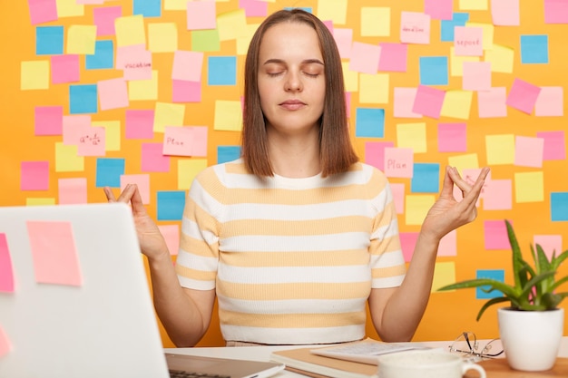 Binnenopname van een ontspannen, mooie blanke vrouw, gekleed in een gestreept T-shirt, poserend tegen een gele muur met kleurrijke memokaarten die yoga beoefenen op de werkplek terwijl ze achter een laptop zitten