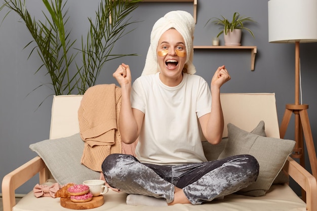 Binnenopname van een opgewonden, extreem gelukkige vrouw met een wit t-shirt in een handdoek op haar hoofd zittend op de bank, haar vuisten gebald en zich verheugend over haar vrije dag poserend met pleisters die cosmetische ingrepen doen