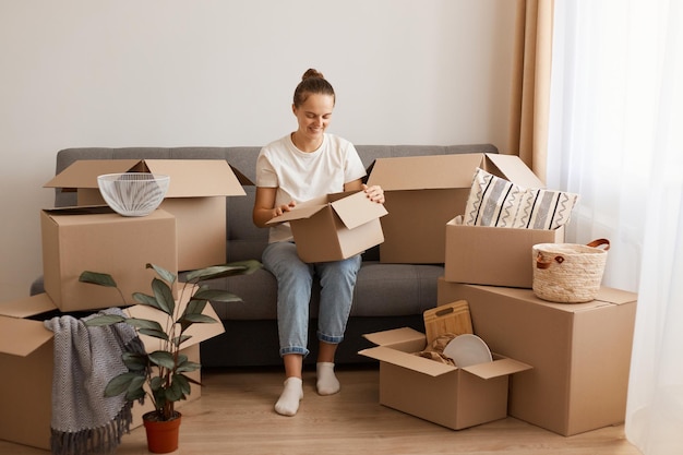 Foto binnenopname van een positief lachende vrouw met een wit t-shirt en een spijkerbroek die op de bank zit in een nieuw huis of appartement na een verhuizing, een kartonnen doos vasthoudt, naar binnen kijkt, pakketten uitpakt.