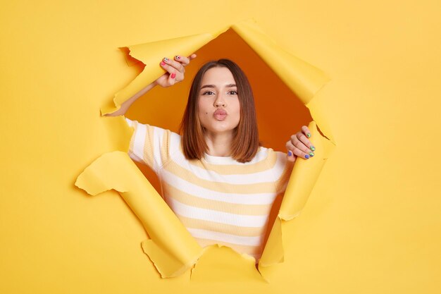 Binnenopname van een romantische, mooie vrouw met een gestreept shirt die luchtkussen naar de camera stuurt en liefde uitdrukt en zacht kijkt door een gat in geel papier