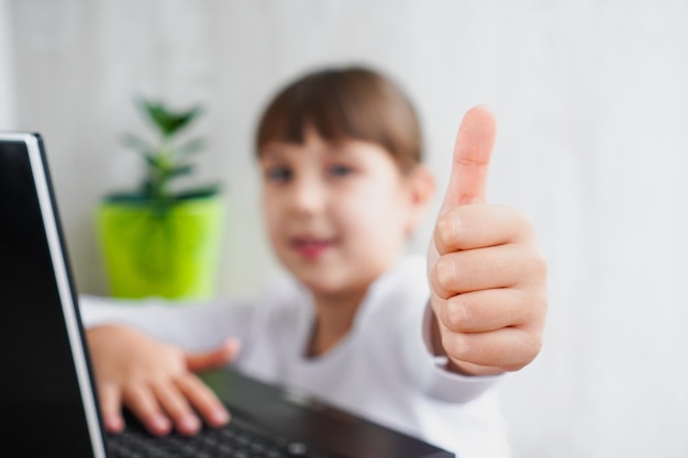 Binnenopname van een schattig donkerharig vrouwelijk kind in een wazige zitting in de buurt van een notebook met duimen omhoog