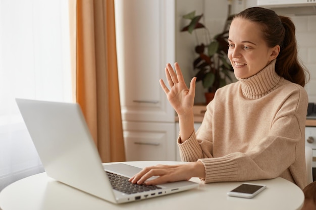Foto binnenopname van een schattige vrouwelijke vrouw met een glimlach met donker haar in een beige trui die in de keuken zit en op een laptop werkt met een online gespreksvideogesprek