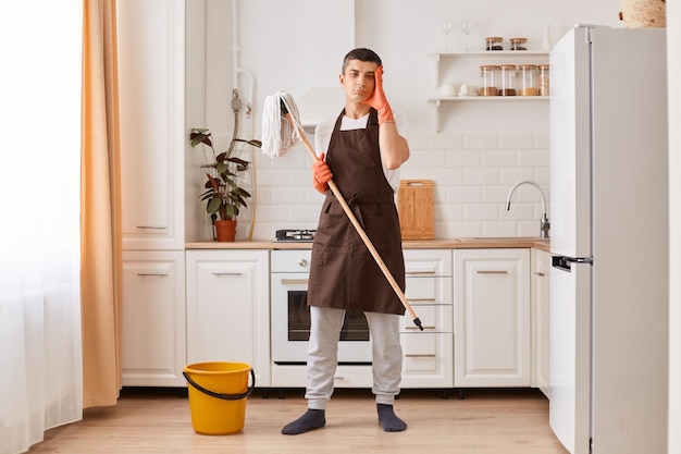 Binnenopname van een uitgeputte vermoeide man met een bruine schort die in de keuken staat na het wassen van de vloer met hoofdpijn die erg moe is en naar de camera kijkt met een droevige gezichtsuitdrukking