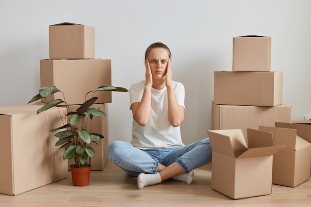 Binnenopname van een uitgeputte vrouw met een wit t-shirt die op de grond zit in de buurt van kartonnen dozen, hoofdpijn heeft, tempels masseert, moe is tijdens het verhuizen naar een nieuw appartement.