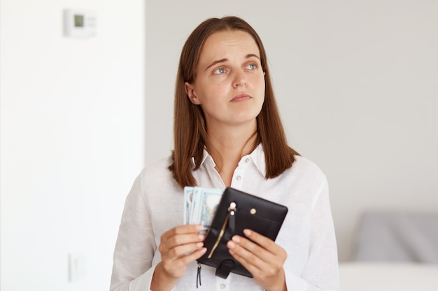 Binnenopname van een verdrietig, overstuur donkerharige vrouw met een wit casual overhemd, een portemonnee met geld in de handen en wegkijkend, uiting gevend aan verdriet, niet genoeg geld hebben om te winkelen.