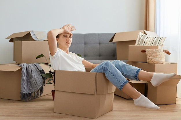 Binnenopname van een vermoeide donkerharige vrouw met een wit t-shirt en een spijkerbroek in een kartonnen doos die naar een nieuw huis verhuist en uitgeput raakt om persoonlijke stapels uit te pakken