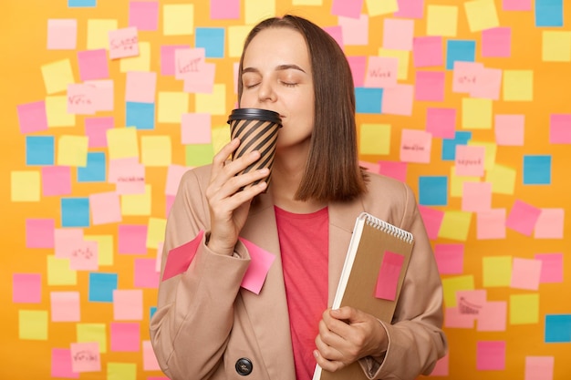 Binnenopname van een vrolijke, ontspannen blanke vrouw in een beige jas die tegen een gele muur poseert met kleurrijke memokaarten die hete koffie ruiken om mee te nemen in een wegwerpbeker