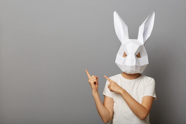 Binnenopname van een vrouw die een wit t-shirt en een papieren konijnenmasker draagt en wijsvingers opzij wijst naar kopieerruimte voor reclame of promotie, geïsoleerd over een grijze achtergrond