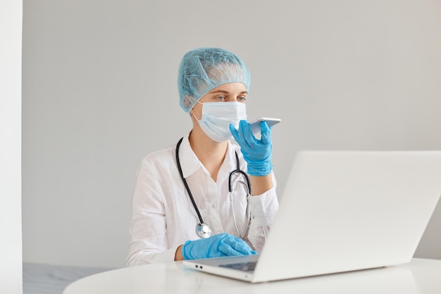 Binnenopname van een vrouwelijke arts die aan tafel zit en op een laptop werkt, met behulp van een mobiele telefoon om spraakberichten of notities op te nemen, met een medische pet, een chirurgisch masker, rubberen handschoenen en een toga.