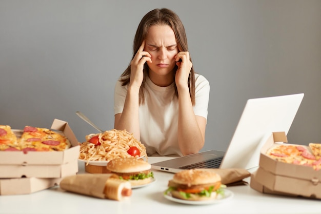 Binnenopname van een zieke, ongezonde vrouw die achter een laptop zit tussen junkfood met vreselijke hoofdpijn, moe en uitgeput geïsoleerd over grijze achtergrond