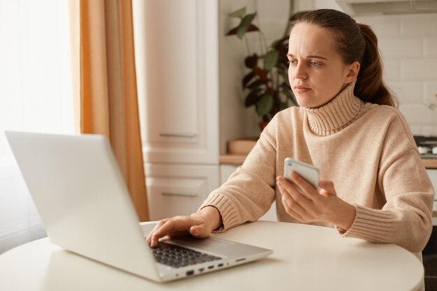 Binnenopname van geconcentreerde hardwerkende blanke vrouw, gekleed in een beige casual stijl trui met paardenstaartkapsel, zittend aan tafel voor een laptopcomputer en met een smartphone in handen.