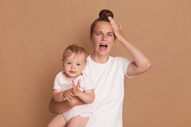 Binnenopname van gestresste depressieve jonge moeder met knotkapsel die haar peuterdochter vasthoudt die schreeuwt dat ze moe en uitgeput is en haar hoofd aanraakt, heeft rust nodig en staat geïsoleerd over bruine achtergrond