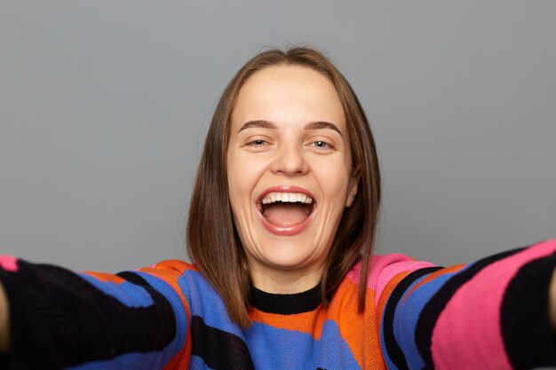 Binnenopname van glimlachende blije opgetogen vrouw die een kleurrijke trui draagt en selfie-standpuntfoto maakt die gelukkig poseert geïsoleerd over grijze achtergrond