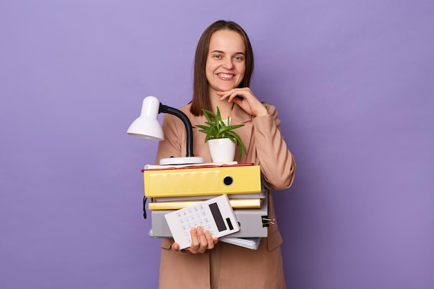 Binnenopname van glimlachende mooie vrouw in een beige jas met veel documentenmappen die naar de camera kijken met een brede glimlach houdt de hand onder de kin poseren geïsoleerd op een paarse achtergrond