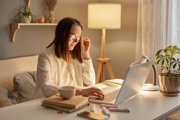 Binnenopname van glimlachende tevreden blanke vrouw met wit overhemd zittend op tafel en werkend op laptop in de buurt van raam met brilmontuur kijkend naar notebookscherm