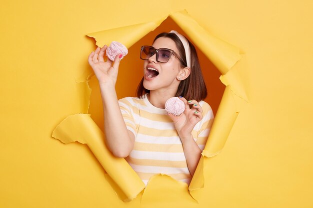 Binnenopname van hongerige vrouw met gestreept shirt en haarband breekt door gele papieren achtergrond terwijl ze marshmallow eet terwijl ze met open mond staat