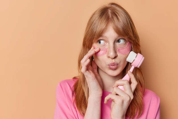 Binnenopname van jong vrouwelijk model met gemberhaar houdt de lippen geconcentreerd weggevouwen maakt gebruik van gezichtsmassager past roze hydrogelpleisters toe onder de ogen poses tegen beige achtergrond lege ruimte