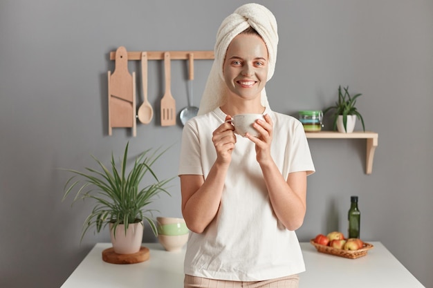 Binnenopname van jonge ontspannen vrouw met cosmetisch masker in badhanddoek op haar hoofd die warme drank, thee of koffie drinkt in de keuken na het nemen van een douchebad thuis schoonheidsbehandeling hydratatie