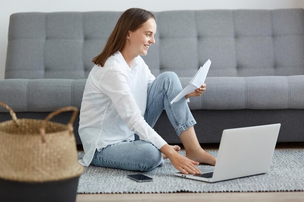 Binnenopname van lachende donkerharige vrouw in wit overhemd en spijkerbroek zittend op de vloer in de buurt van de bank met behulp van laptop en met papieren document betalende openbare nutsbedrijven