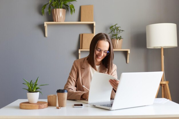 Binnenopname van lachende jonge volwassen mooie bruinharige vrouw met beige jas poserend op kantoor werkend op laptop en en met papieren documenten genietend van haar werk