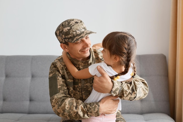 Binnenopname van lachende man soldaat met camouflage-uniform en pet poserend met zijn dochter die elkaar omhelst en blij is om te zien na lange scheiding